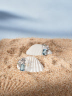 Aquamarine gemstone stud earrings beside a white seashell, floral halo with clear stones
